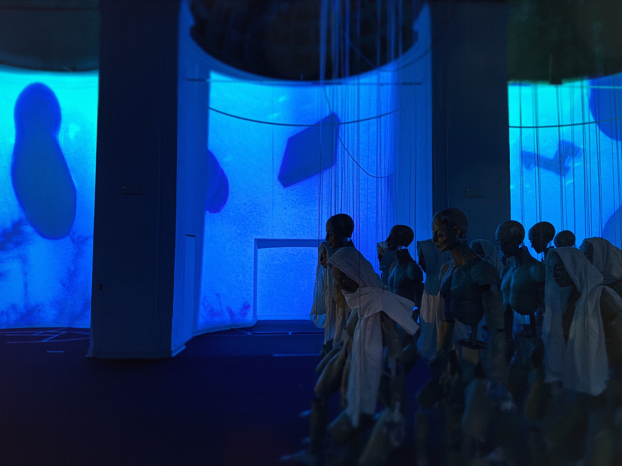 Figure sospese come marionette illuminate da proiezioni blu nell'installazione AIDA